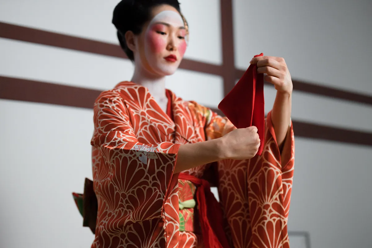 A performer in kimono and traditional white stage makeup holding a dance cloth, representative image